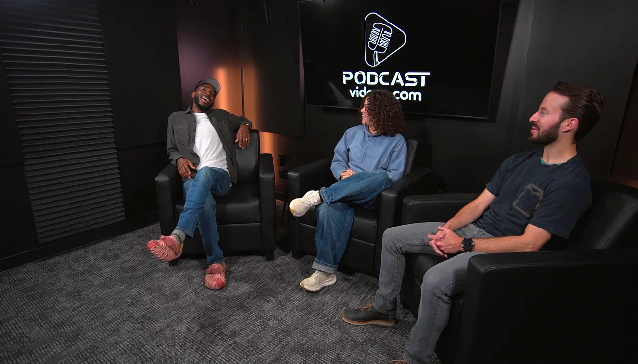 Three people sit in a dark podcast studio, smiling and engaged in conversation. The backdrop features a large screen with a podcast logo.