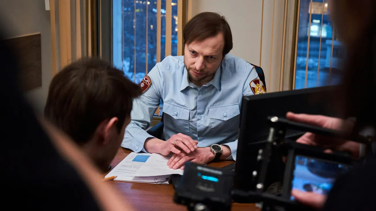 Man in uniform sits at a desk during a tense conversation, papers scattered before him. Filming equipment is visible in the foreground. Mood is serious.