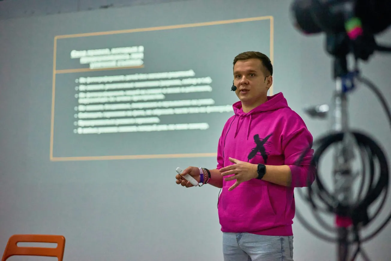 A person in a bright pink hoodie speaks confidently, holding a clicker in front of a blurred presentation slide.