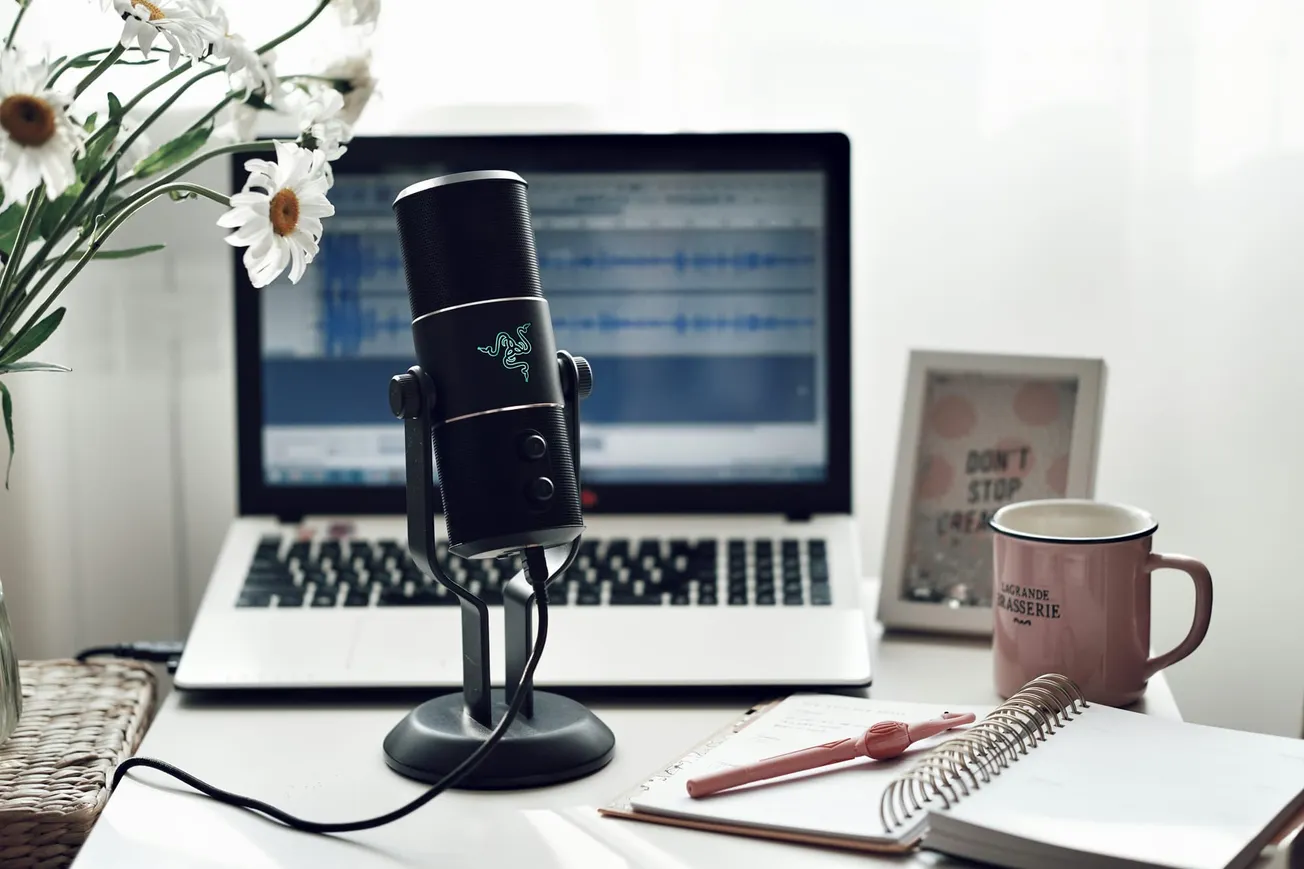 A workspace with a microphone, open laptop showing audio editing software, notebook, pen, mug, flowers, and an inspirational framed quote. Cozy and creative.