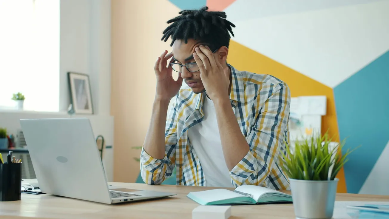 A man sits at a desk in a colorful office, holding his head and looking frustrated. An open laptop, notebook, and potted plant are on the desk.