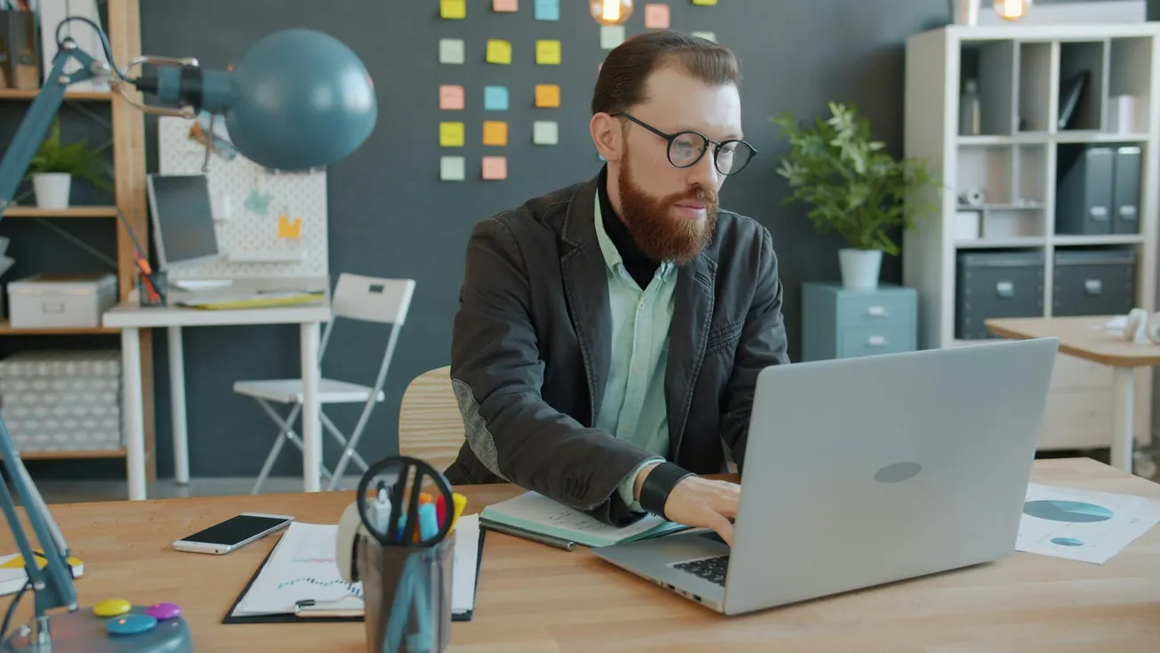 A bearded man with glasses is focused on a laptop in a modern office. A bulletin board with sticky notes is behind him, creating a productive atmosphere.