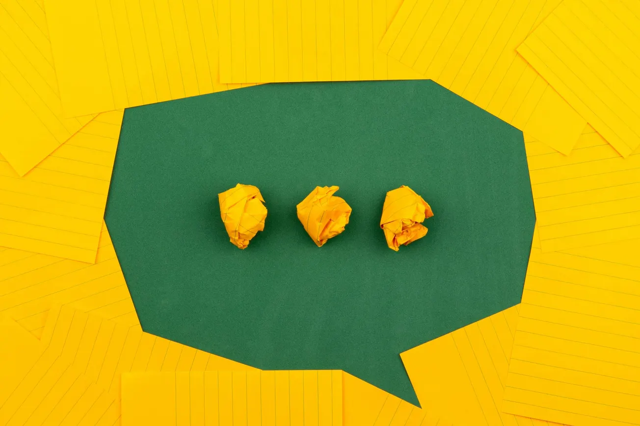 A green speech bubble on a background of scattered yellow lined paper, with three crumpled yellow balls inside, suggesting communication or ideas.
