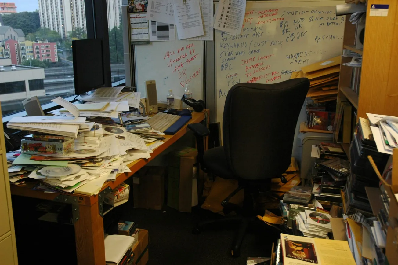 A cluttered office with papers and CDs stacked on a wooden desk. A black chair faces a messy whiteboard with notes. Large window shows cityscape.