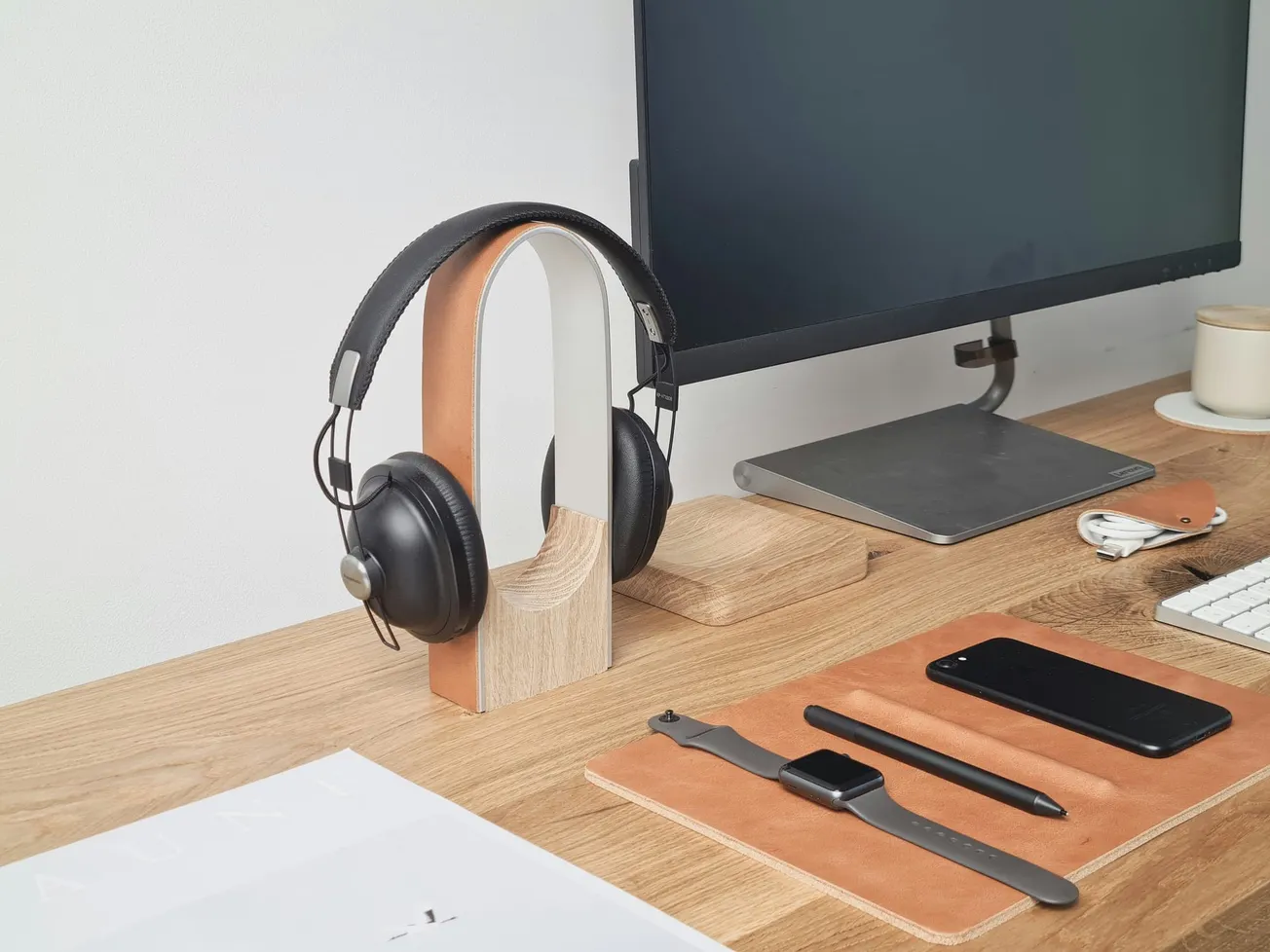 A tidy workspace with a wooden desk, featuring headphones on a stand, a monitor, smartphone, smartwatch, stylus on a pad, keyboard, and a coffee cup.