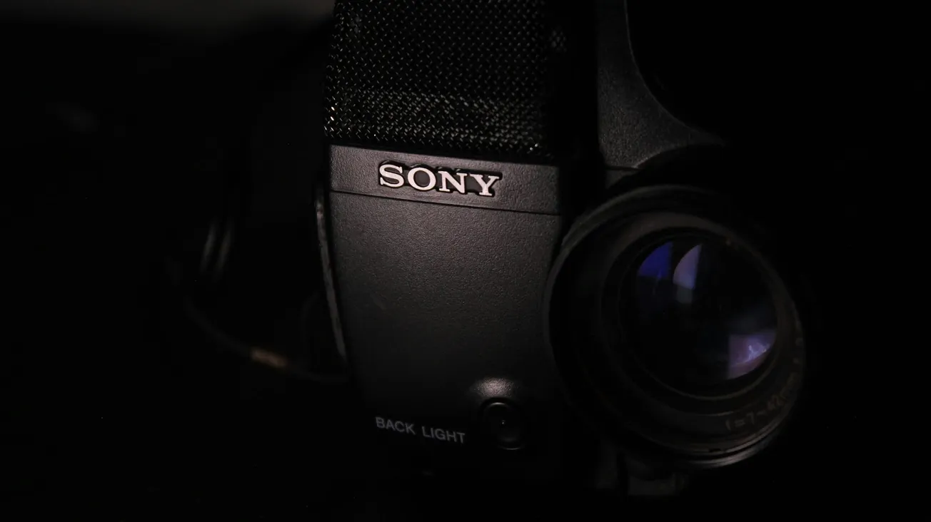 Close-up of a Sony video camera in low light; the lens reflects blue hues, creating a mysterious and focused atmosphere; "Sony" logo is visible.
