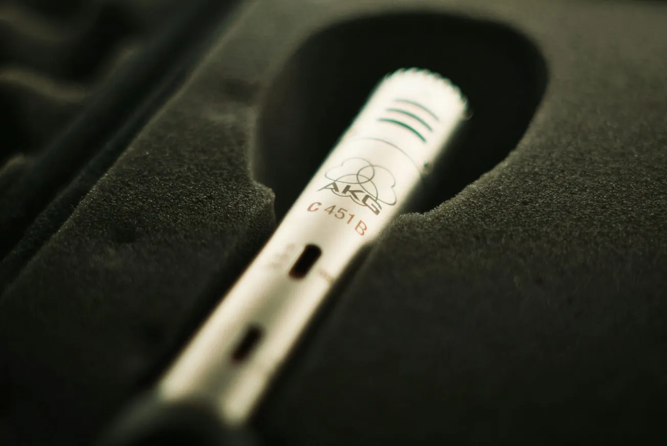 Close-up of a silver microphone labeled "AKG C 451 B" nestled in a dark foam case, conveying a sense of professional audio equipment ready for use.