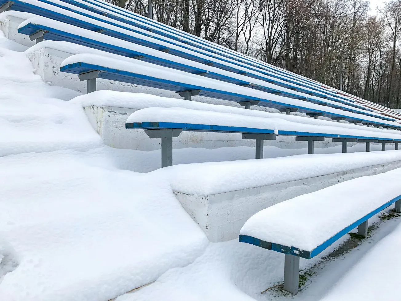 Snow-covered bleachers in an outdoor stadium create a serene winter scene. Blue seats contrast with the white snow. Bare trees stand in the background.
