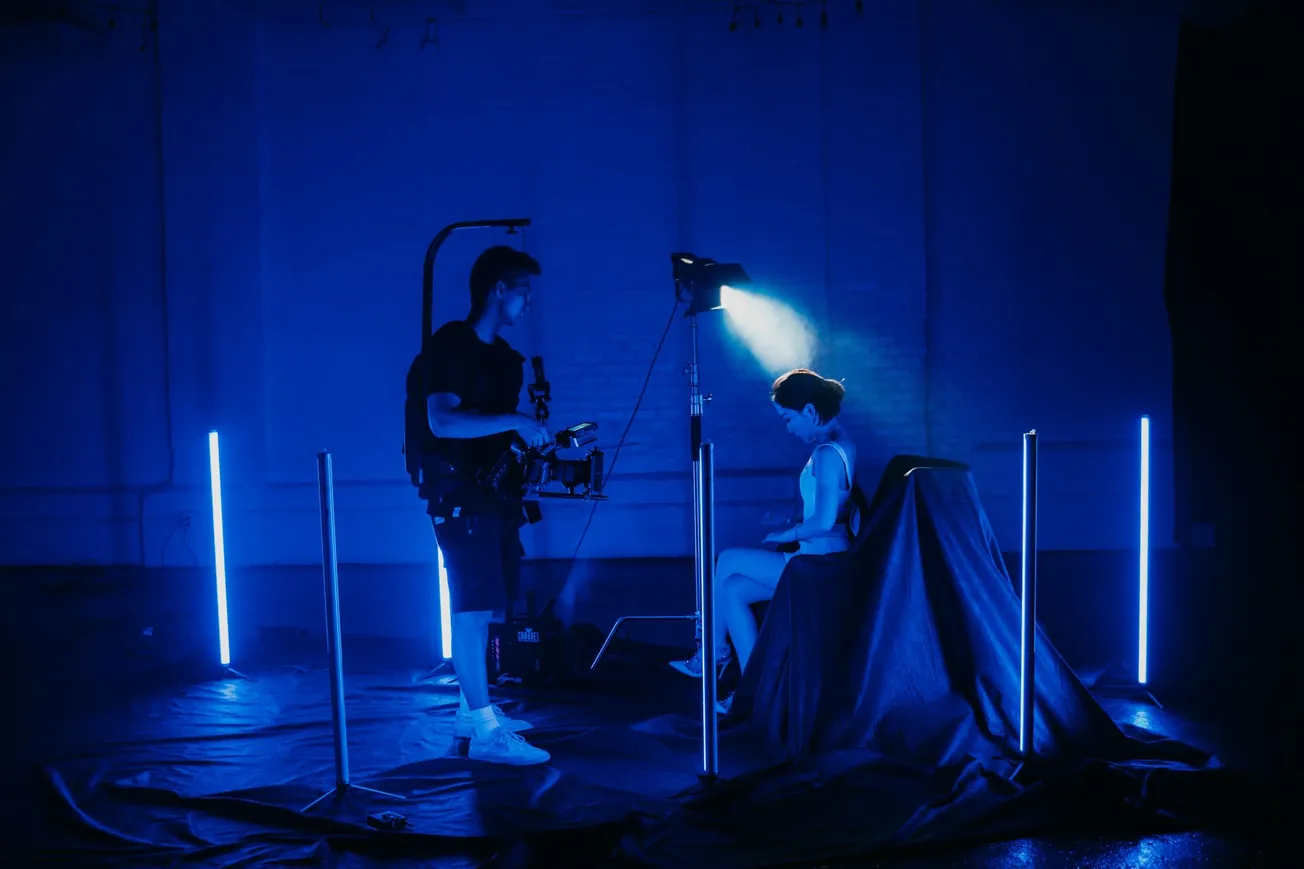 A dimly lit studio with blue lighting shows a person sitting in a chair under a spotlight, as a camera operator stands nearby, creating a mysterious mood.