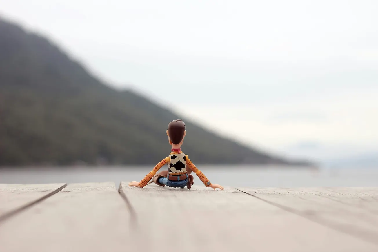 Toy figure in cowboy attire sits on a wooden surface, gazing out at a misty, mountainous landscape beside a serene body of water, evoking nostalgia.
