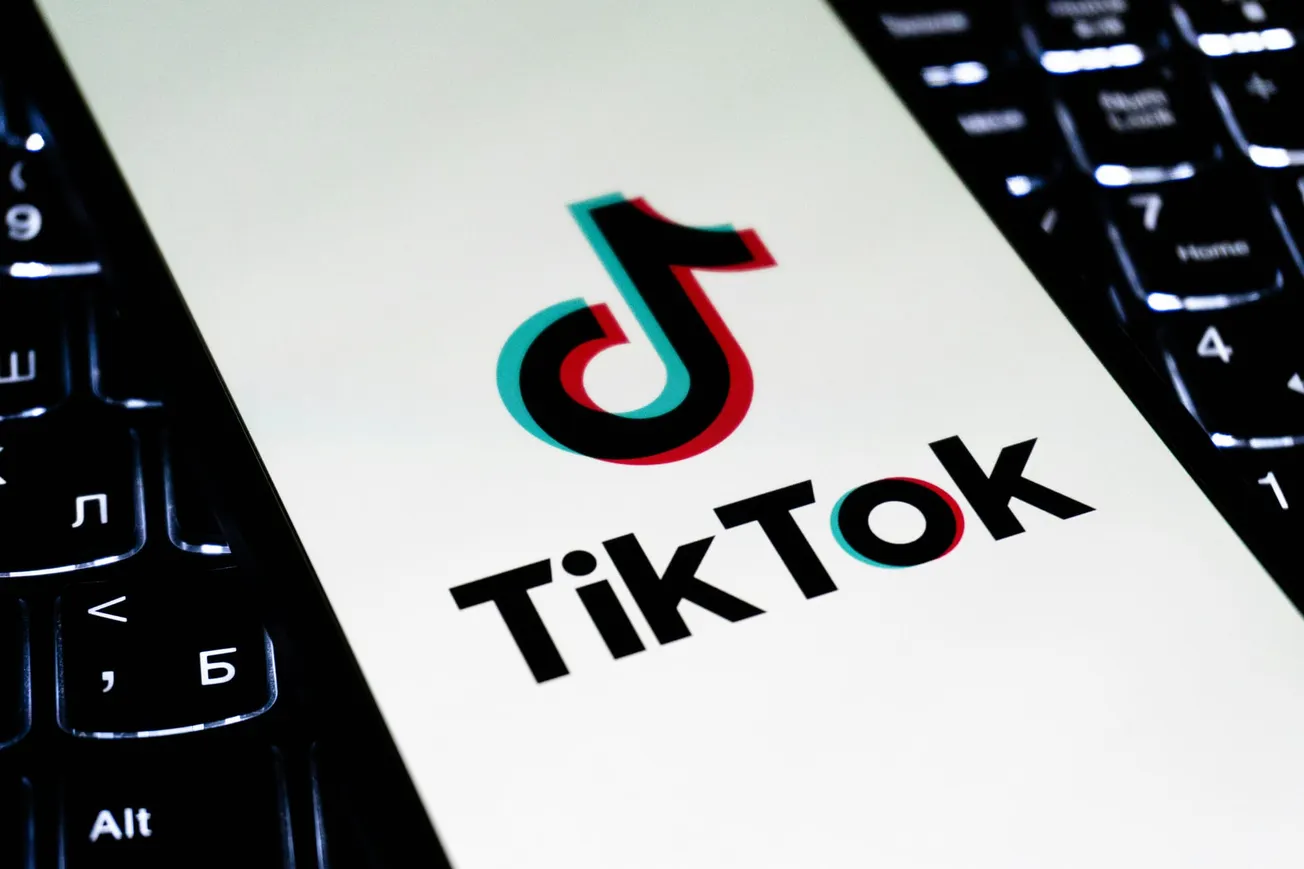 Smartphone screen displays the TikTok logo, a musical note icon, over a white background. The device rests on a lit keyboard, symbolizing technology.