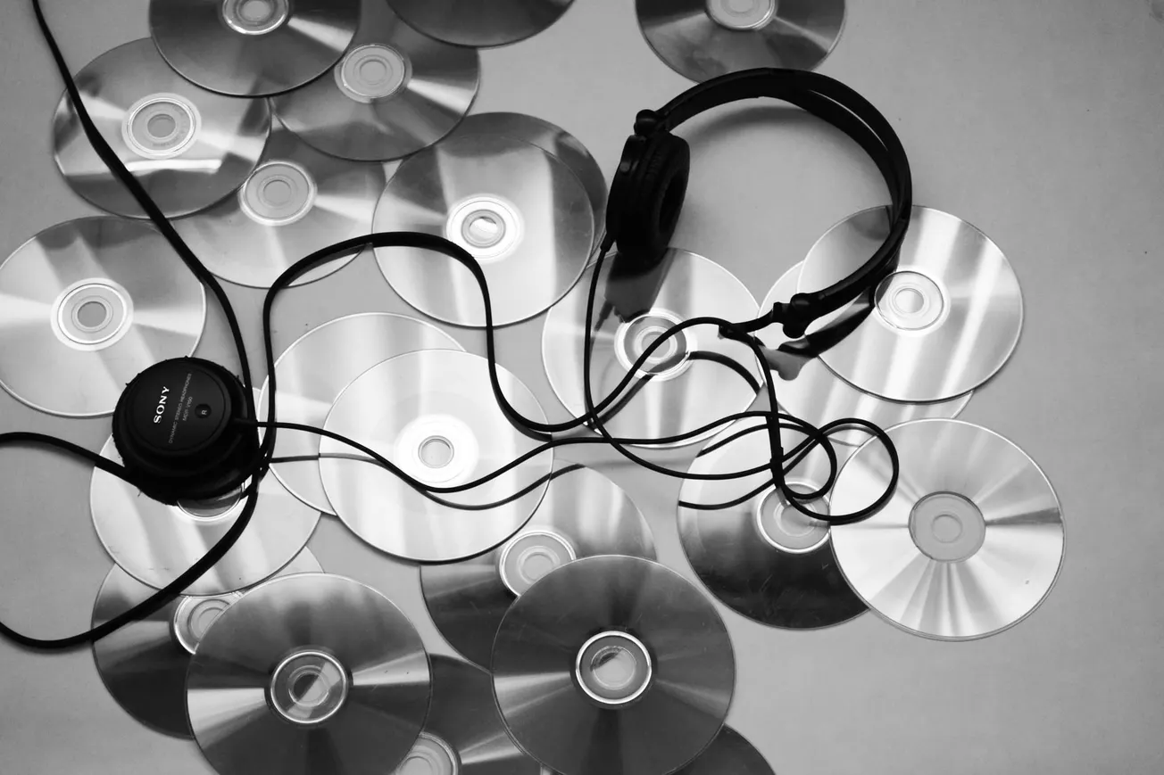 Scattered CDs with reflective surfaces surround black headphones, which have a tangled cord, on a flat surface. The tone is nostalgic and retro.