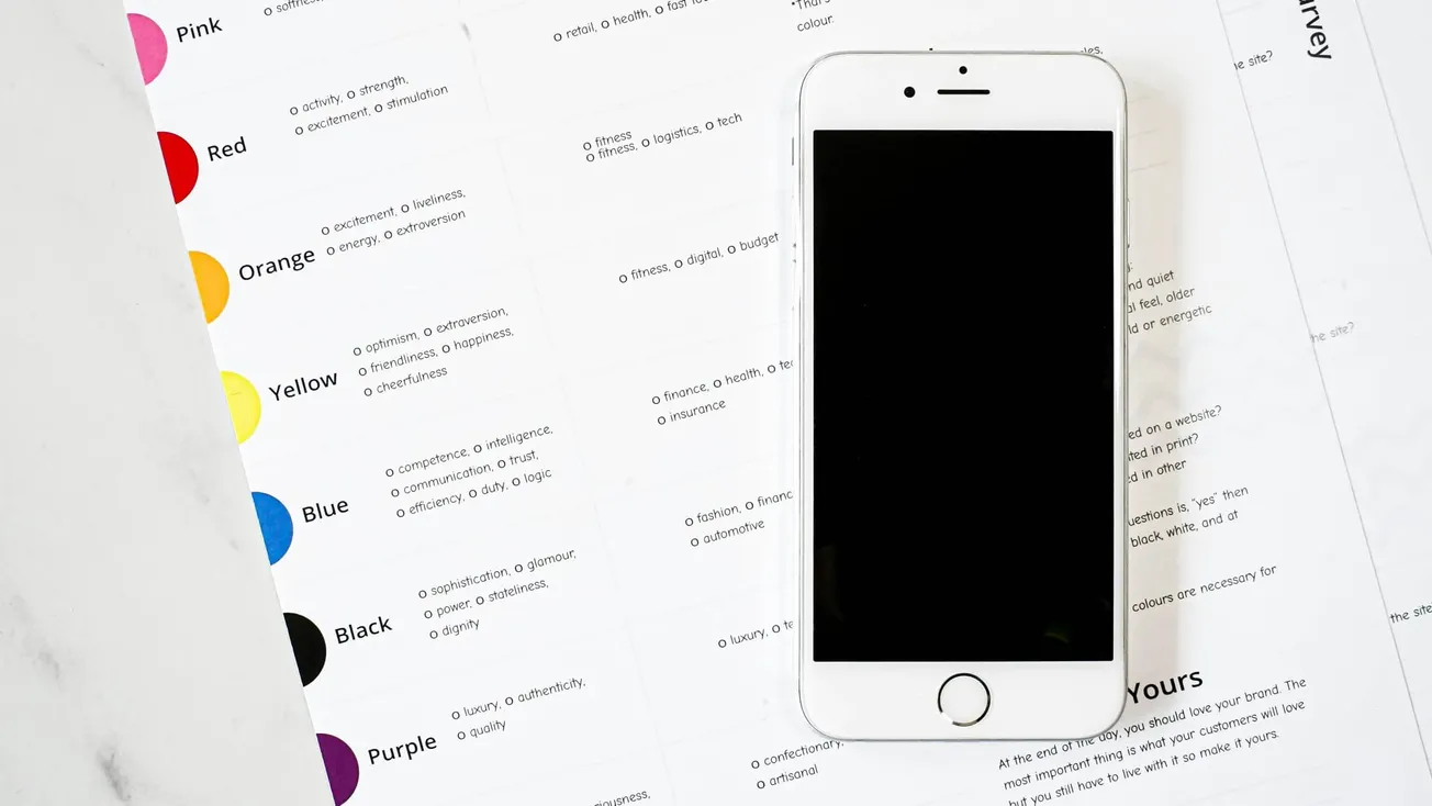 A smartphone with a blank screen is placed on a spread of printed pages featuring color swatches and descriptions. The setting is bright and minimalistic.