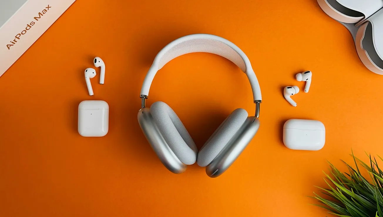 Silver over-ear headphones are centered on an orange background, surrounded by two pairs of white earbuds in their cases. A small plant adds a touch of greenery.