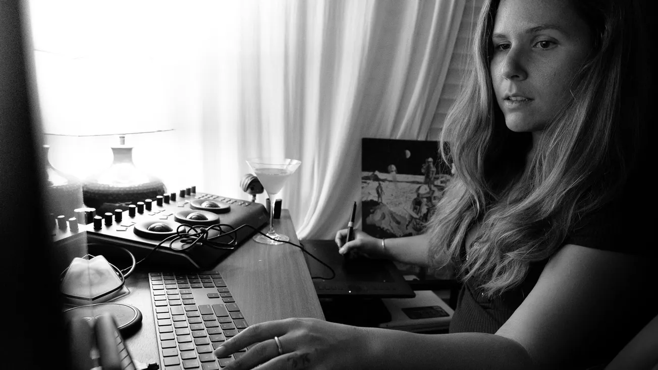 A woman works intensely at a computer in a dimly lit room. The monochrome setting features a keyboard, drawing tablet, and a lamp, creating a focused atmosphere.