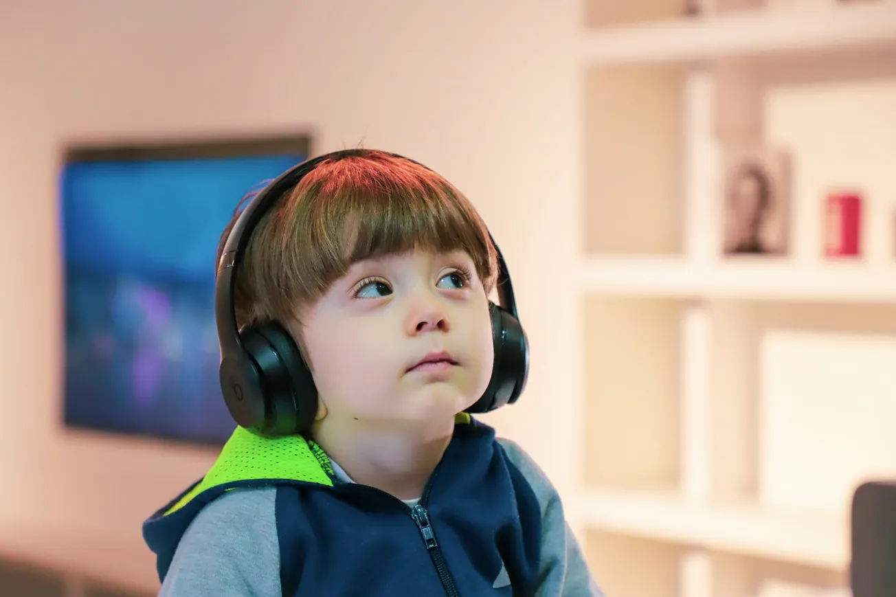 A young child wearing large headphones gazes upward, appearing thoughtful. The background is a softly lit room with a blurry TV screen and shelves.