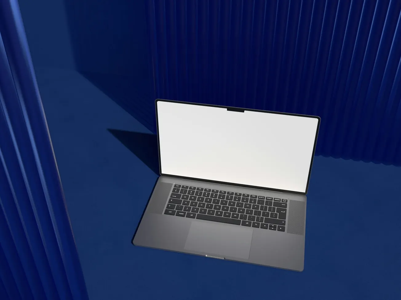 A sleek gray laptop with a blank white screen sits on a dark blue surface against a backdrop of blue corrugated pillars.