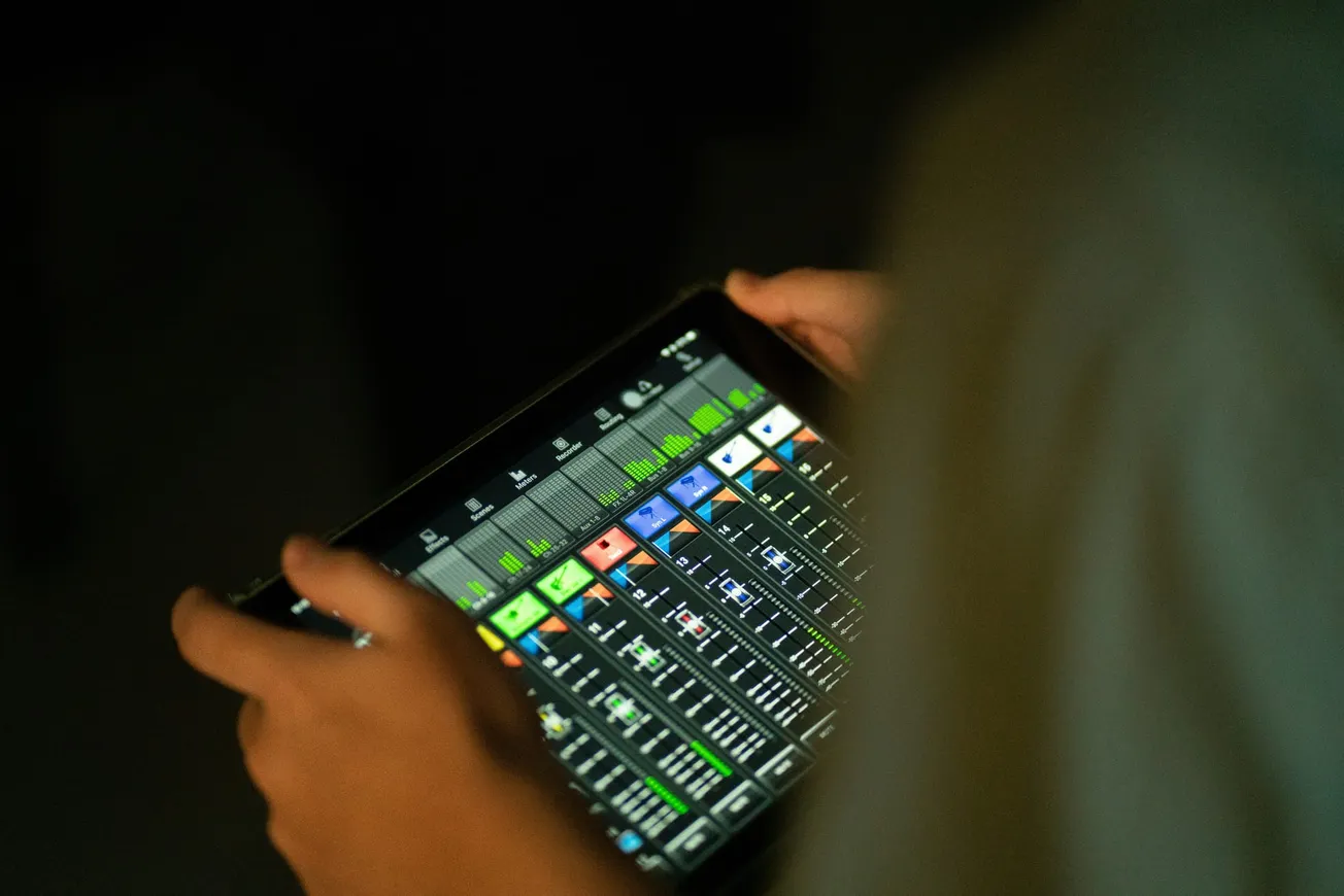 Hands holding a tablet displaying a colorful digital audio mixer interface with sliders and levels. The ambient lighting is dim, creating a focused mood.