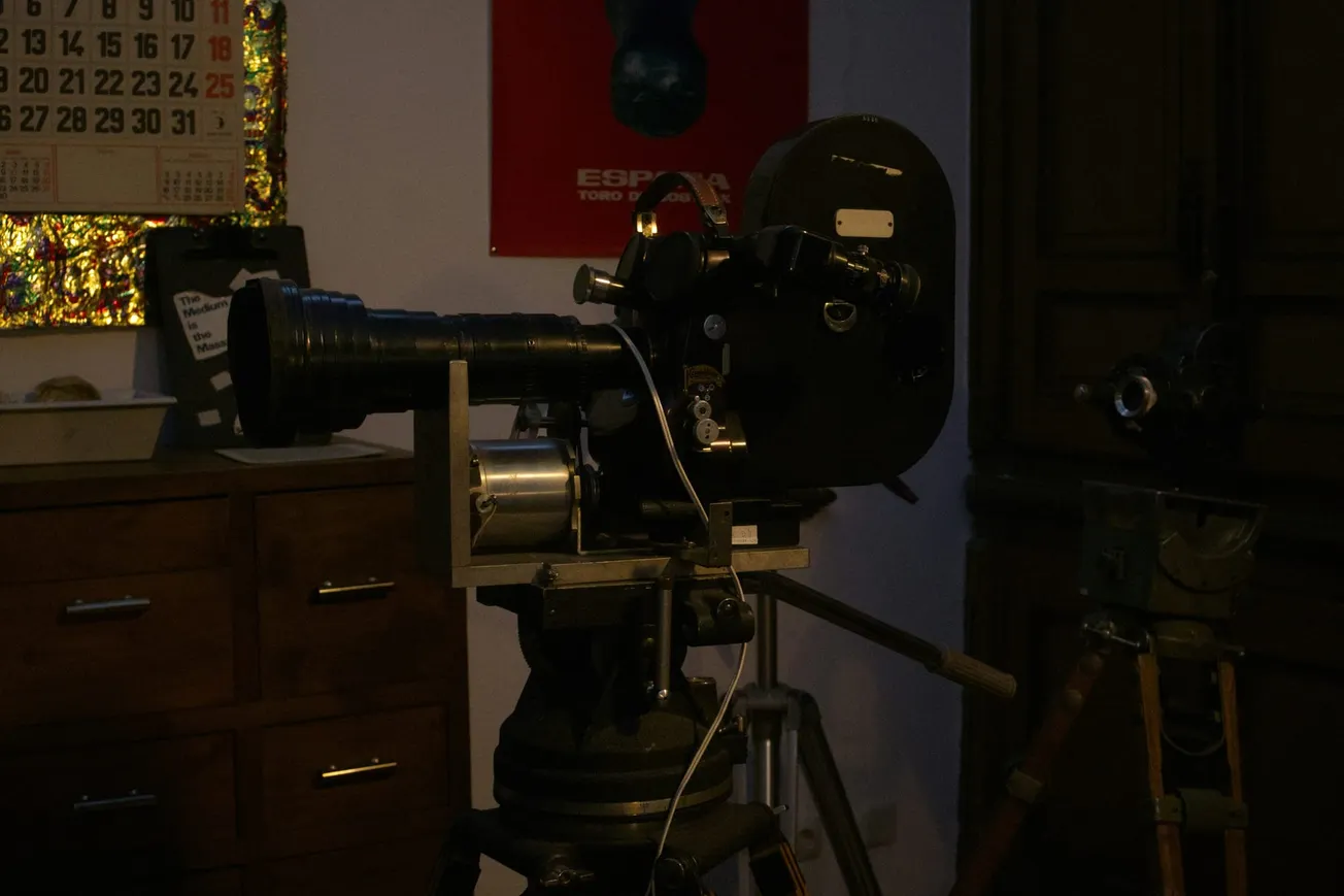 Dimly lit room with a vintage film camera on a tripod in the foreground. A calendar and red poster are visible on the wall, creating a nostalgic tone.