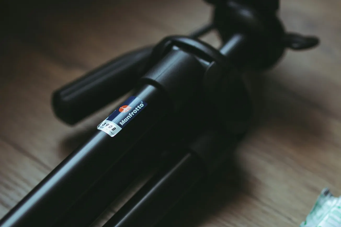 A black Manfrotto tripod resting on a wooden floor, showcasing its legs and a brand label. The lighting is dim, contributing to a moody atmosphere.
