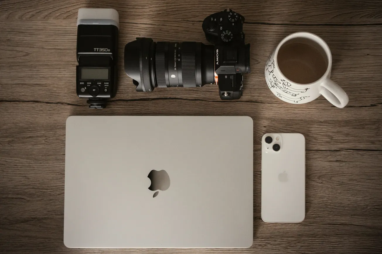 A flat lay image on a wooden surface featuring a laptop, camera, flash, smartphone, and a mug of coffee. The mood is organized and professional.