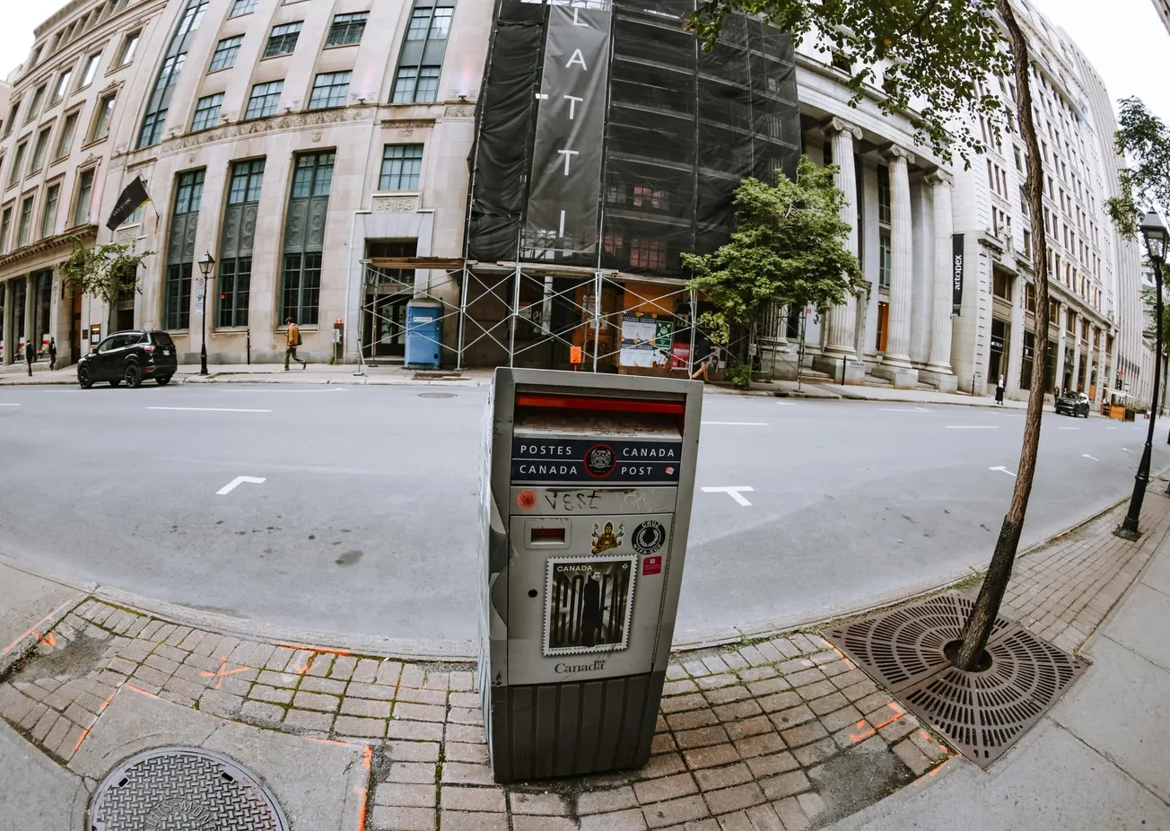 A Canada Post mailbox is in the foreground on a city street. Behind it, there are large historic buildings, one under renovation. A few cars and people are visible.