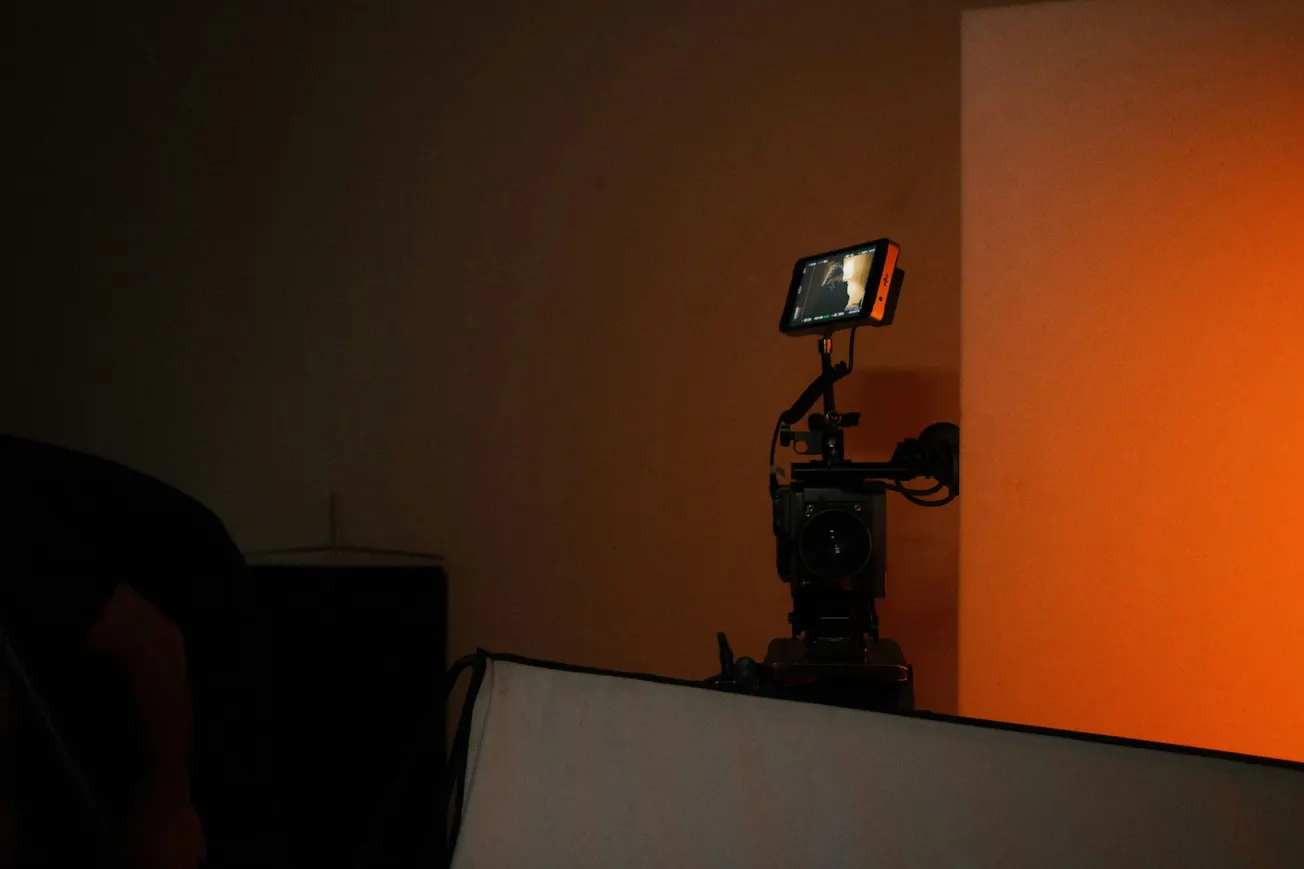 A professional video camera setup is positioned against a warm orange backdrop. It features a small monitor displaying a blurred image, creating a cinematic ambiance.