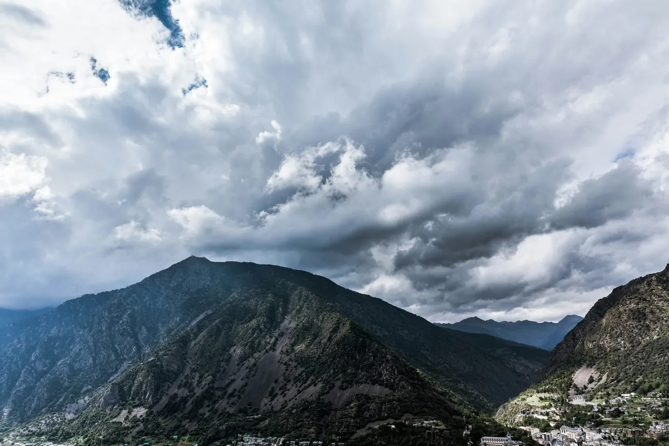 Dramatic view of a steep mountain range under a cloudy, overcast sky, evoking a sense of impending storm. A small village nestles in the valley below.