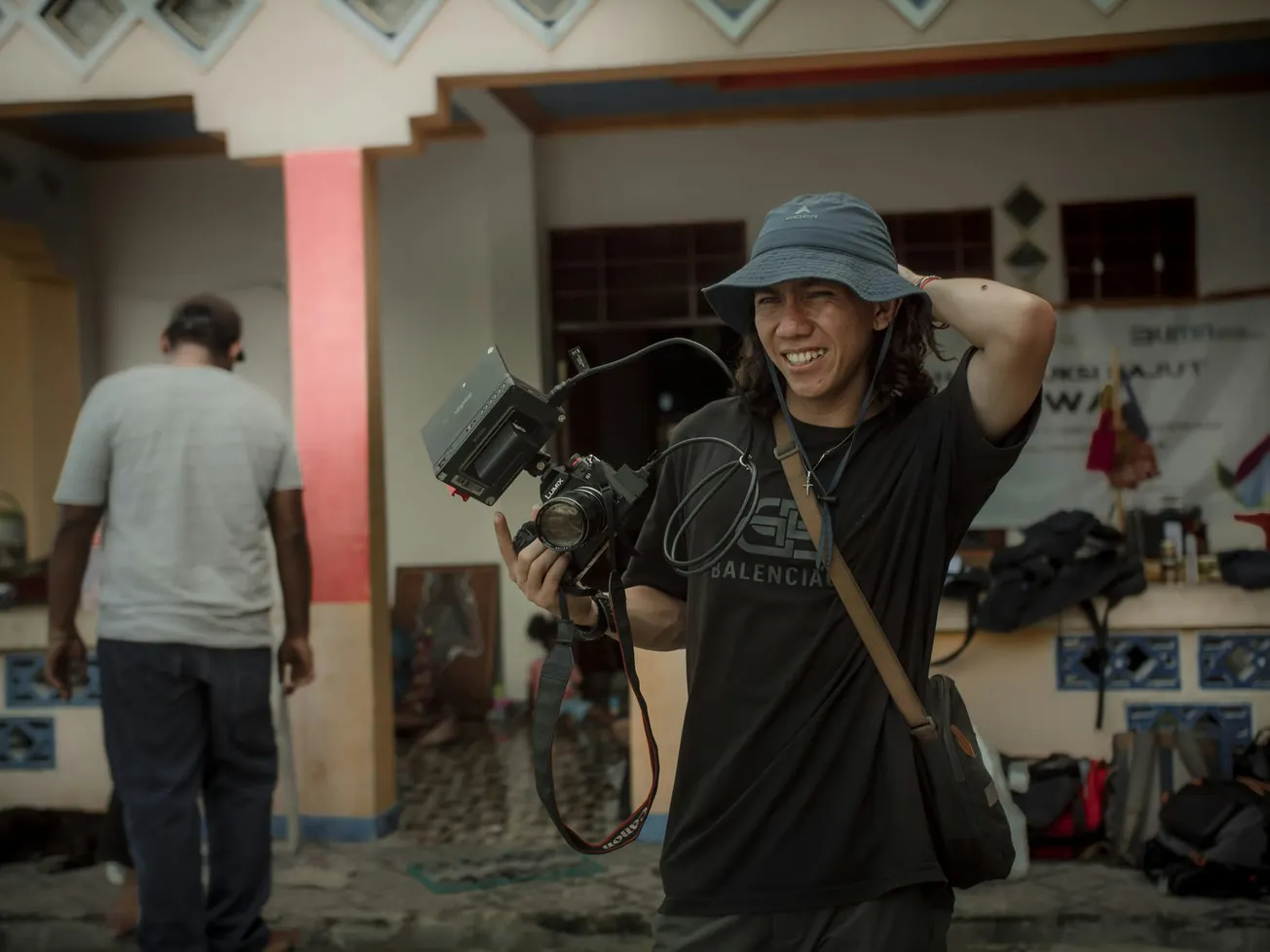 A person smiling and holding a camera, wearing a blue bucket hat and casual attire, standing outside with gear in the background, exuding a joyful and relaxed vibe.