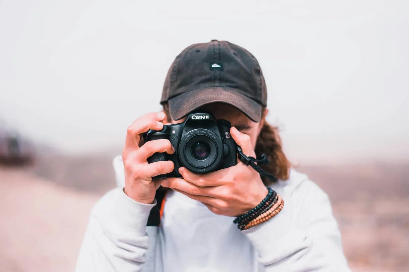 A person in a cap, focused on taking a photo with a Canon camera. They're dressed casually, with layer bracelets, against a blurred outdoor backdrop.