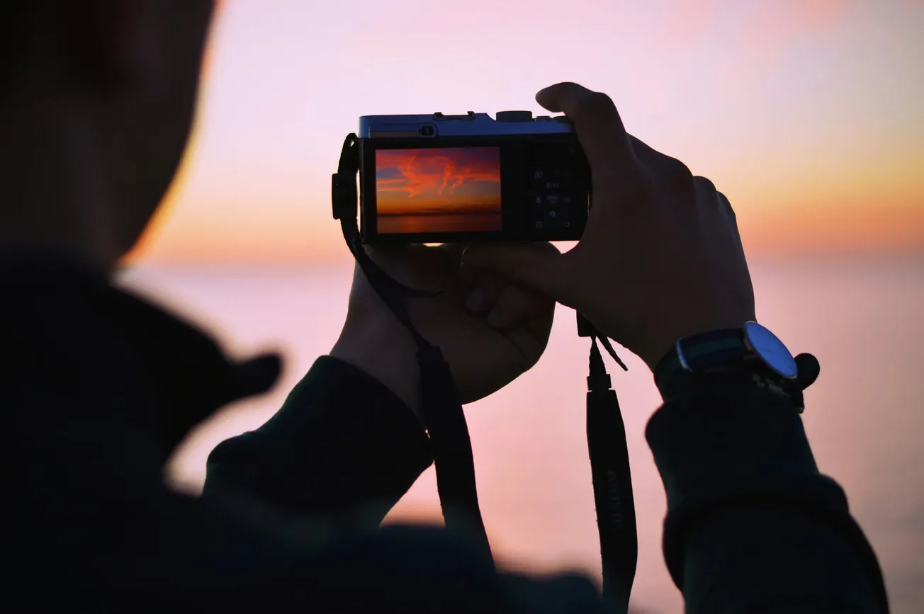 A person holds a camera capturing a vivid sunset over the ocean. The scene reflects vibrant orange and pink hues, conveying serenity and focus.
