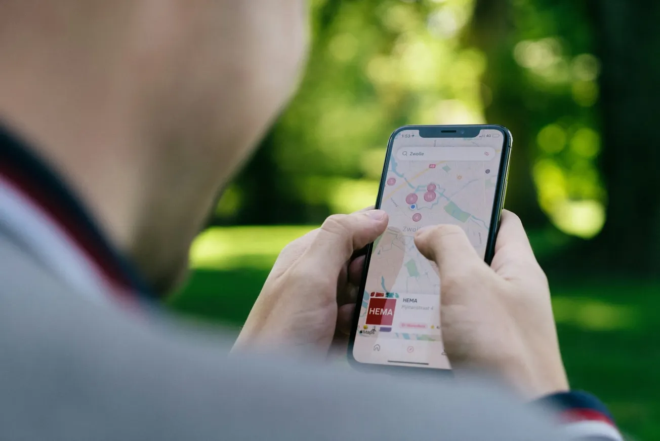 A person uses a smartphone to navigate a map app outdoors. The screen shows location points on a map. Bright greenery blurs in the background.