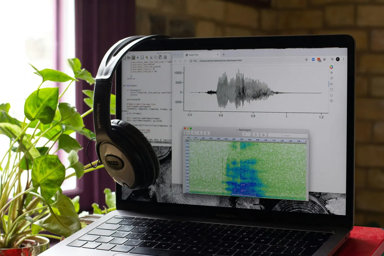 A laptop with code and waveform visuals on screen, draped with headphones, sits by a potted plant. The setup suggests audio analysis work.