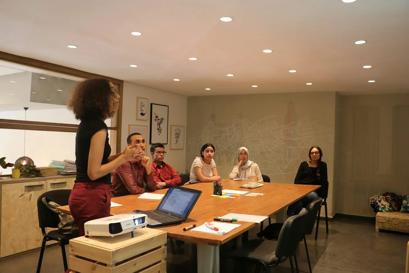 A person stands presenting to five attentive individuals seated around a wooden table in a modern conference room. A laptop and projector are in use.