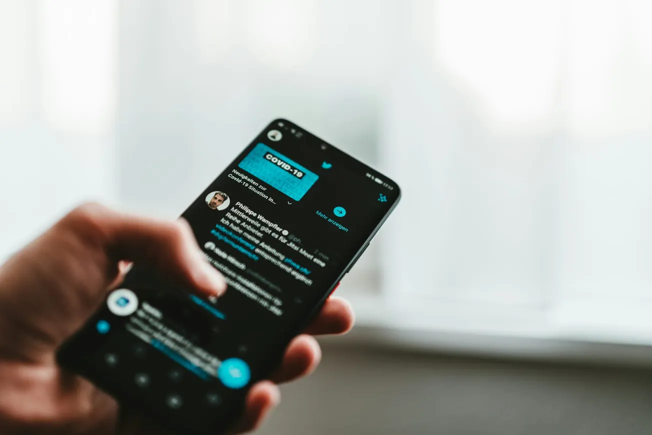 A hand holds a smartphone displaying a Twitter feed focused on COVID-19. The background is softly blurred, conveying a calm and focused tone.