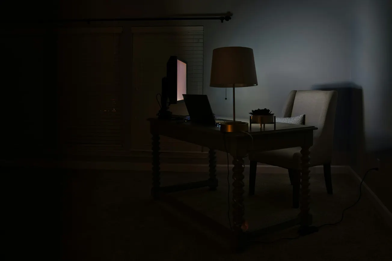 A dimly lit room features a computer on a desk with a glowing screen. Beside the desk is a chair and a lamp, creating a quiet, contemplative atmosphere.