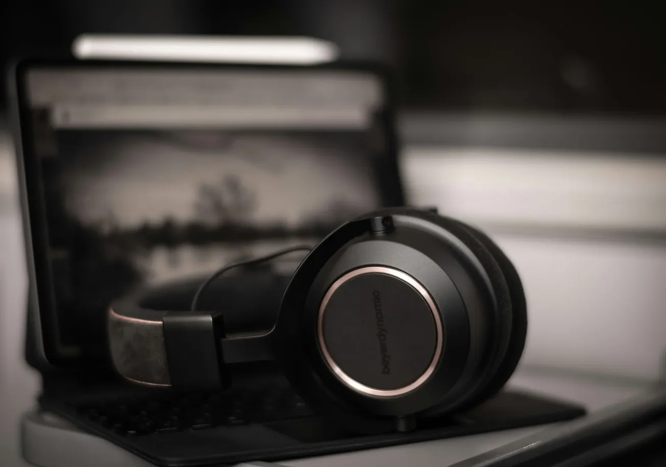 Close-up of large black headphones resting on a laptop keyboard, with the screen showing a blurred grayscale image. The scene is dimly lit, creating a moody atmosphere.