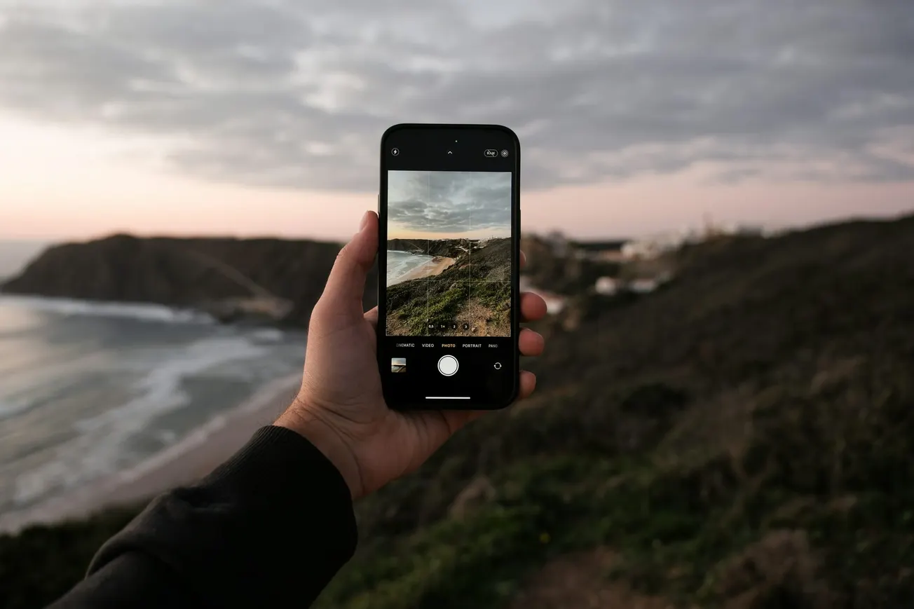 A hand holds a smartphone capturing a scenic coastline with cliffs and ocean waves. The sky is overcast, revealing a tranquil, muted sunset.