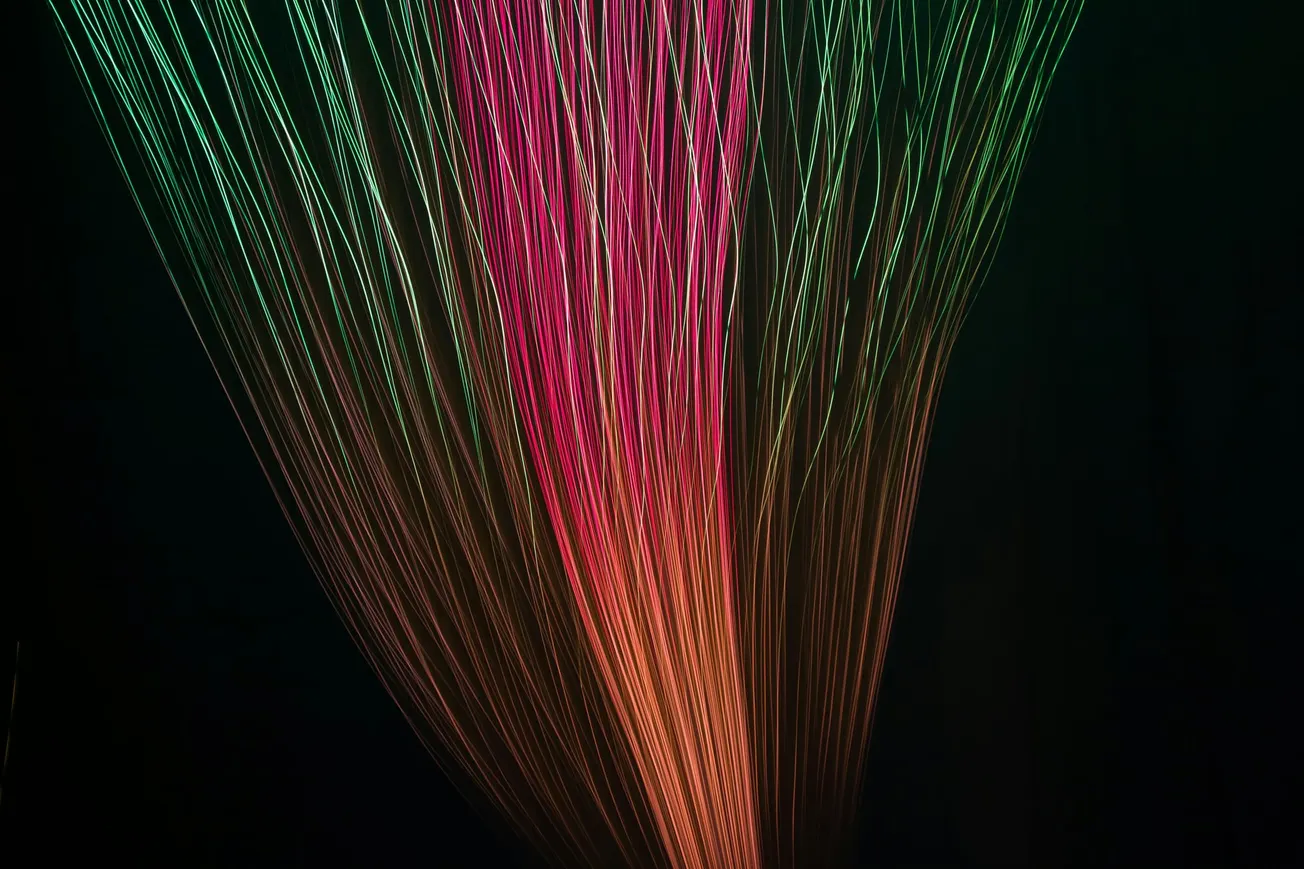 Vibrant light trails in green, pink, and orange converge against a dark background, creating a dynamic, ethereal, and mesmerizing visual pattern.