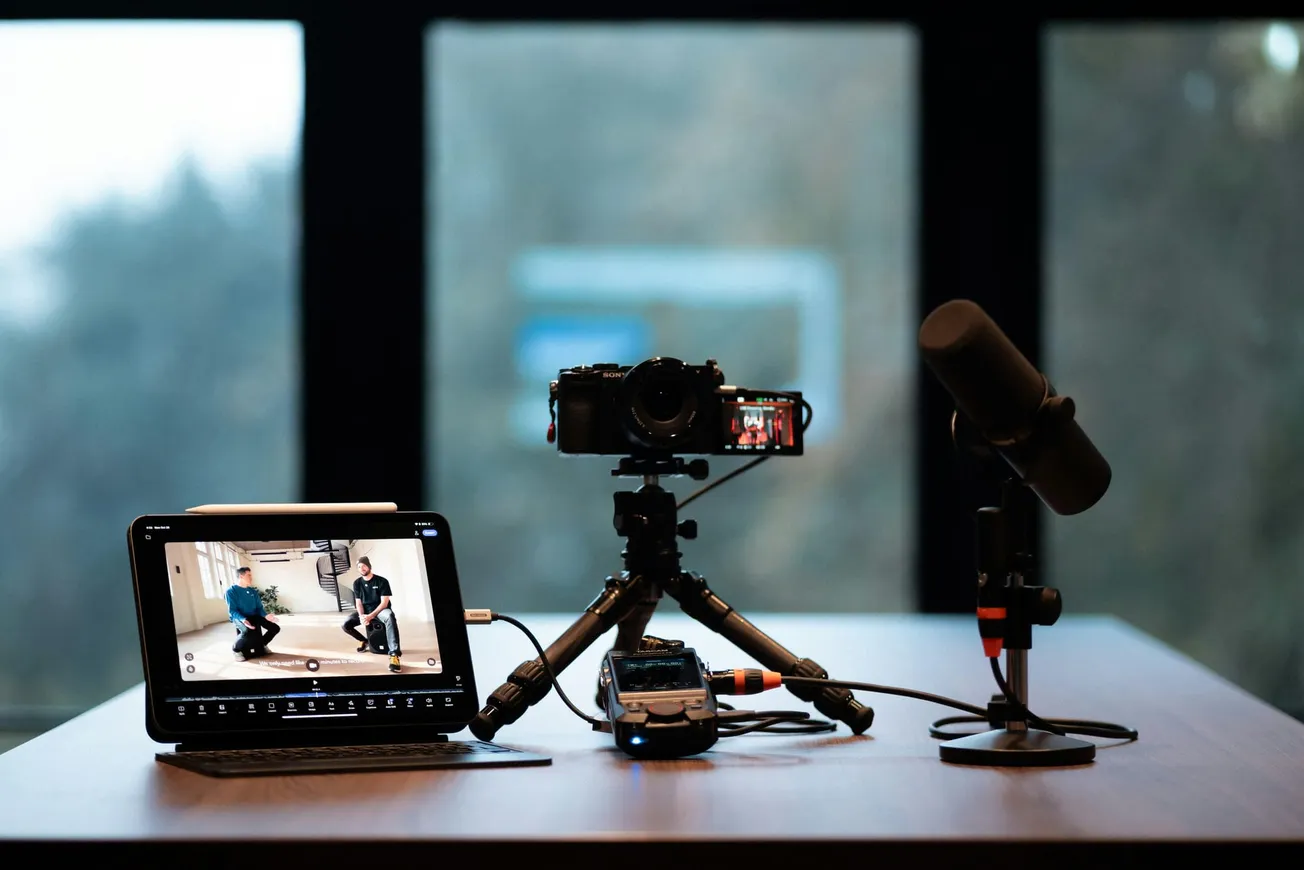 A podcast setup on a table includes a camera on a tripod, a tablet displaying two people talking, and a microphone. A large window is in the background.