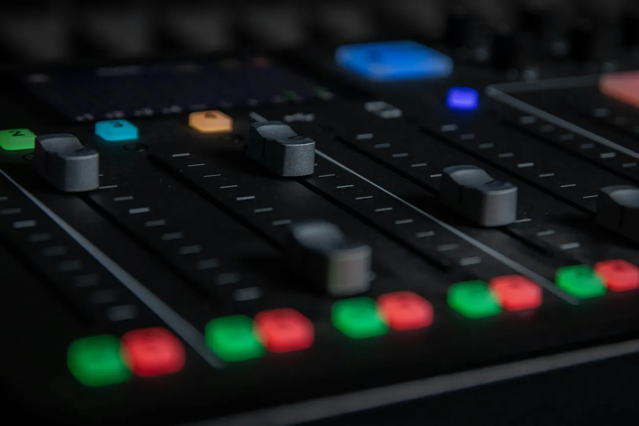 Close-up of a sound mixing board with sliders and colorful buttons. Dim lighting gives a focused, professional studio atmosphere.