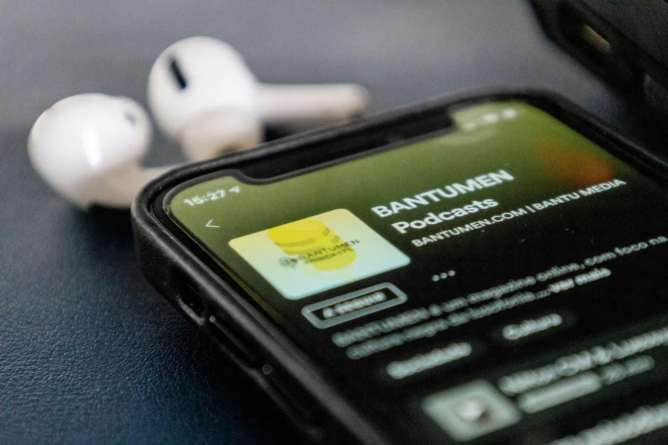 Smartphone displaying a podcast app with "BANTUMEN Podcasts" onscreen. Nearby, two white wireless earbuds rest on a dark surface.
