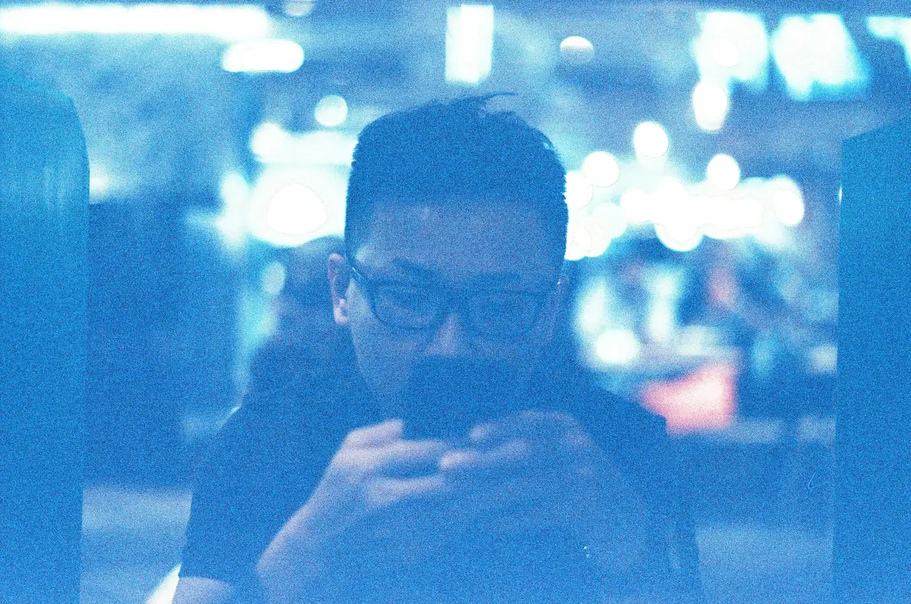 A man with glasses is focused on his phone in a dimly lit, blurry setting with an intense blue tint. The background displays bokeh lights, creating a dreamy ambiance.