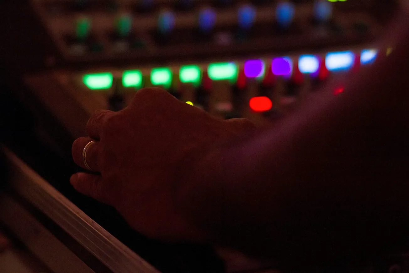A hand adjusts controls on an audio mixing board, surrounded by colorful, softly glowing lights. The scene is dimly lit, conveying focus and concentration.