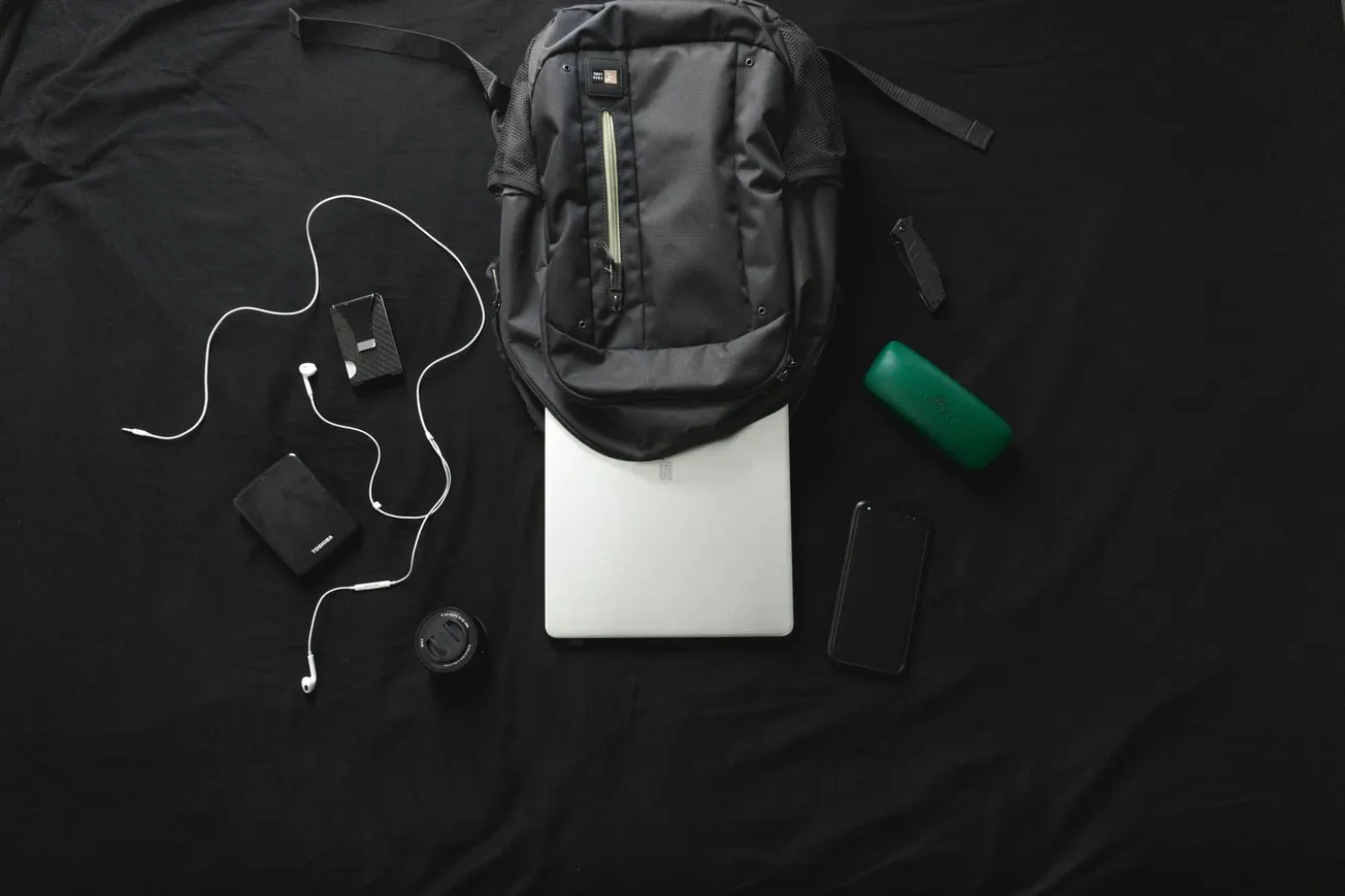 A backpack, laptop, phone, earbuds, wallet, portable speaker, watch, and a camera lens are neatly arranged on a black background.