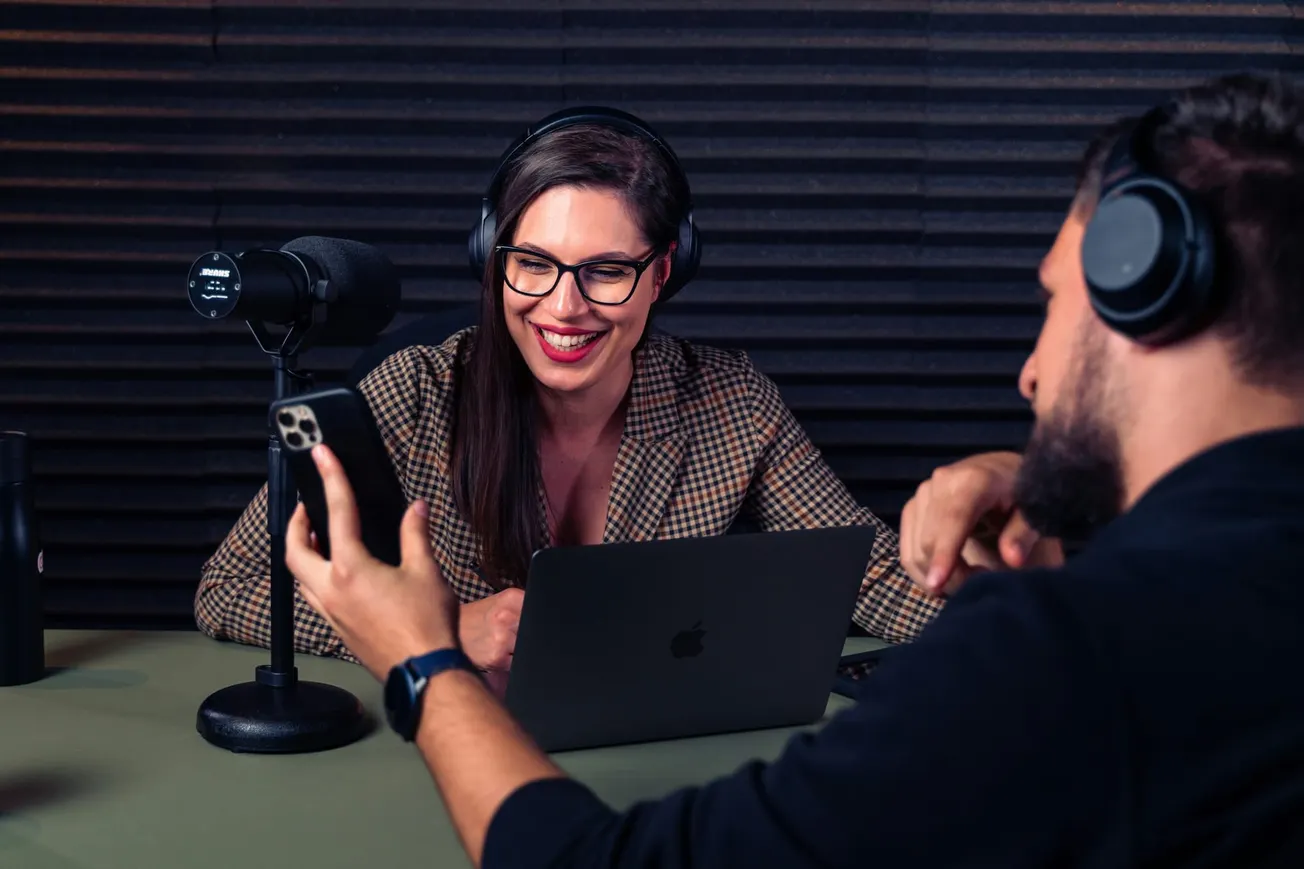 A woman in glasses and headphones smiles in a podcast studio with a microphone and laptop. A man in headphones holds a phone, creating a collaborative vibe.