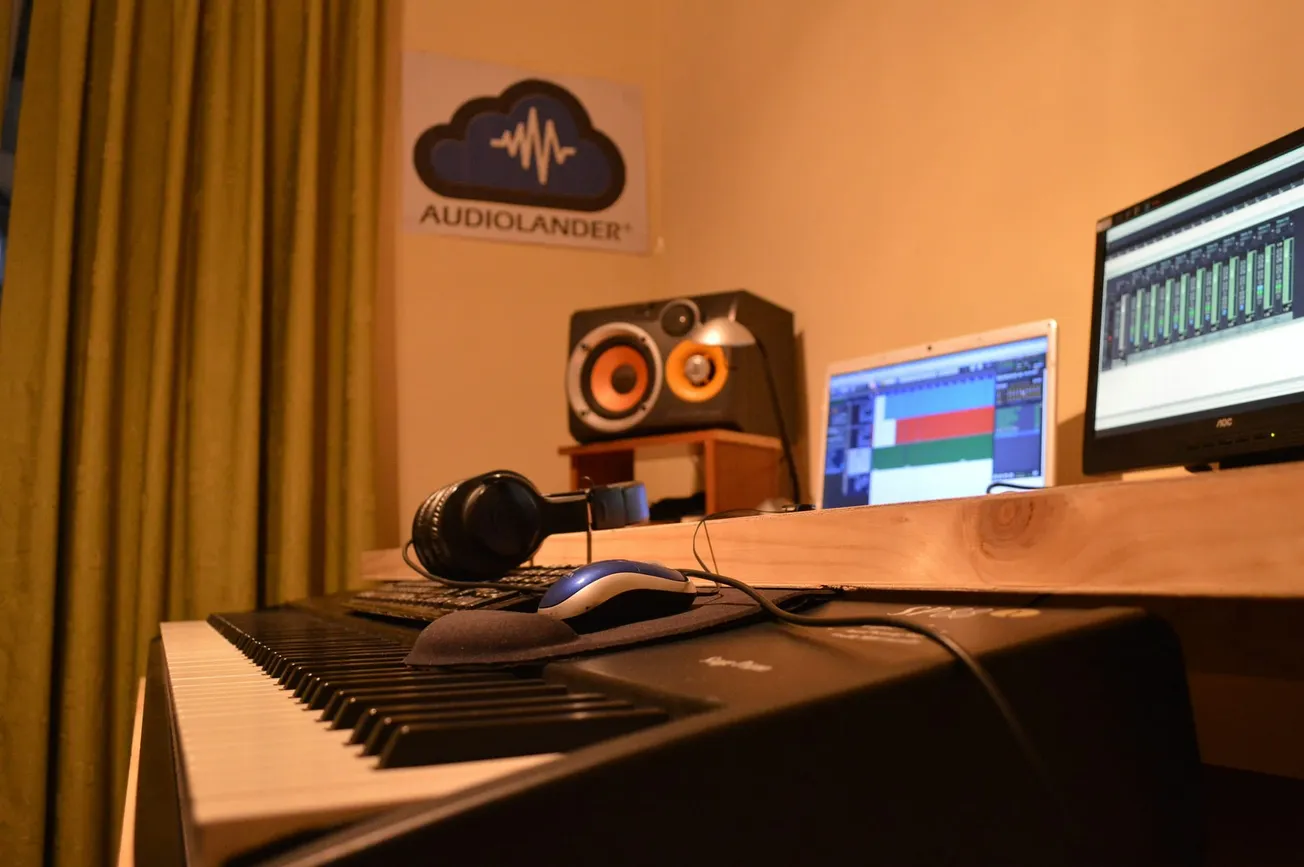 A cozy music studio setup featuring a keyboard, headphones, and computer monitors displaying audio editing software. Speakers and a poster enhance the creative ambiance.