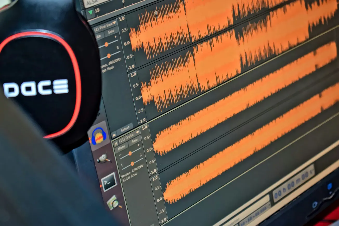 A close-up view of a computer monitor displaying orange sound waves in an audio editing software, with a pair of "Pace" headphones partially visible in the foreground.