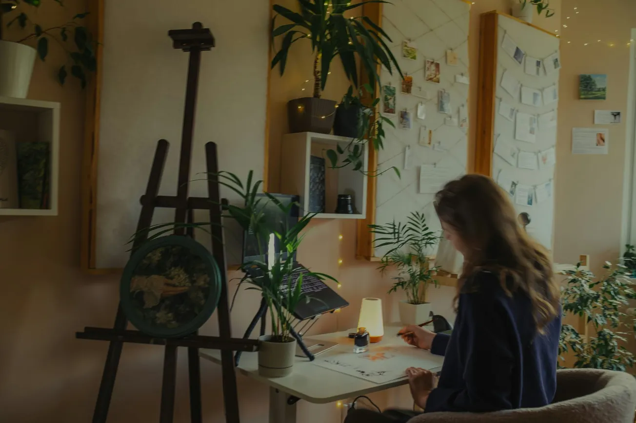 Cozy art studio with a woman drawing at a desk. Surrounded by plants, artwork on an easel, and softly lit with string lights, creating a calm ambiance.