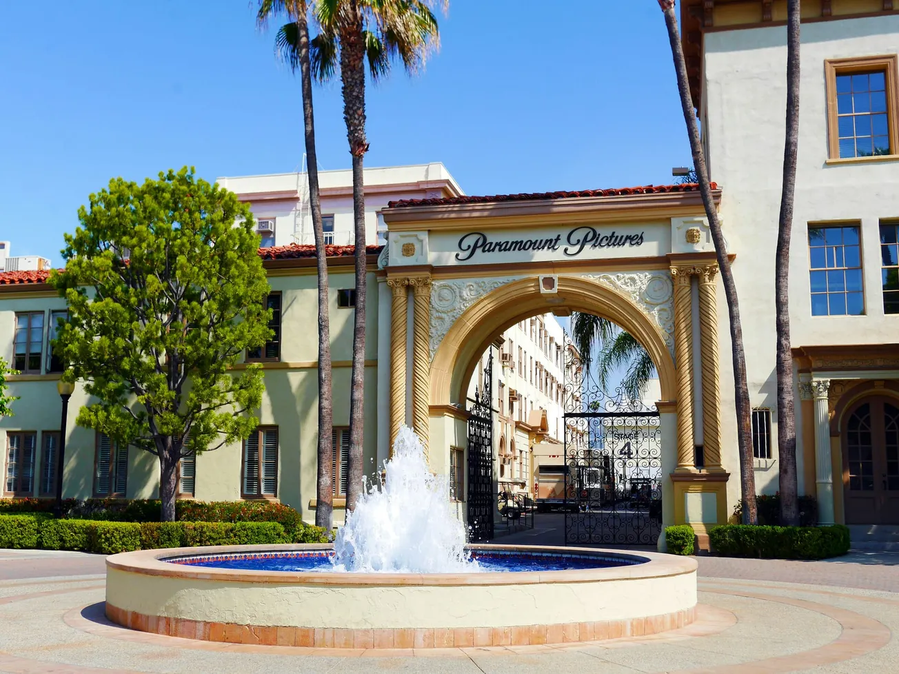 A large circular water fountain sits in the foreground of the ornate Paramount Pictures studio gate, framed by palm trees and Mediterranean-style buildings.